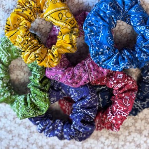 May include: Six colorful scrunchies made from fabric with a bandana print. The scrunchies are in shades of green, yellow, blue, pink, red, and purple.