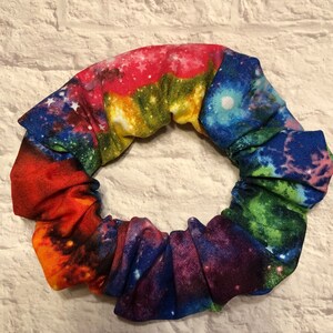 May include: A colorful galaxy print scrunchie with a rainbow of colors, including red, orange, yellow, green, blue, and purple. The scrunchie is made of a soft, stretchy fabric and is perfect for holding hair in place.