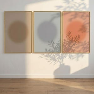 Puede incluir: Tres impresiones artísticas abstractas enmarcadas. Cada impresión presenta un diseño circular suave en tonos beige, gris y coral. Las impresiones están enmarcadas en dorado y están montadas en una pared blanca. La sombra de una planta se proyecta sobre la pared y las impresiones.