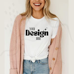 Puede incluir: Una camiseta blanca con el texto "YOUR Design HERE" en negro, combinada con vaqueros azules y una rebeca rosa claro. La rebeca tiene botones grandes y mangas largas. La modelo sonríe.