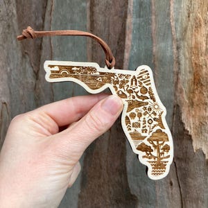 May include: Wooden ornament in the shape of Florida, featuring etched illustrations of palm trees, a lighthouse, and other Florida landmarks. The ornament has a brown leather loop for hanging.