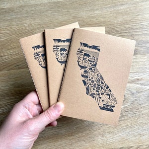 May include: Three brown notebooks with a black outline of the state of California filled with icons representing different aspects of the state. The icons include a bear, a peace sign, a palm tree, a surfboard, a camper, a lighthouse, a mountain, a fish, and a cactus.