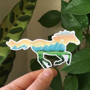 May include: A sticker in the shape of a running horse. The horse is filled with a landscape design featuring mountains, trees, and a sunset. The sticker is held in front of a background of green plants.