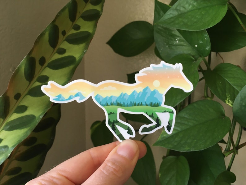 May include: A sticker in the shape of a running horse. The horse is filled with a landscape design featuring mountains, trees, and a sunset. The sticker is held in front of a background of green plants.