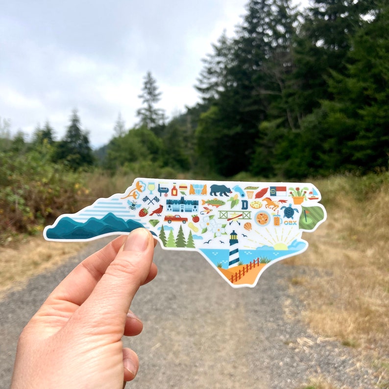Pu&ograve; includere: Un adesivo bianco a forma dello stato della Carolina del Nord con illustrazioni colorate di vari monumenti, attivit&agrave; e simboli che rappresentano lo stato. L'adesivo &egrave; tenuto in una mano su uno sfondo di alberi verdi e un sentiero sterrato.