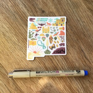 Op de afbeelding: Een sticker met een kleurrijke omtrek van de staat New Mexico met verschillende pictogrammen die de cultuur en het landschap van de staat vertegenwoordigen. De pictogrammen omvatten een luchtballon, een chilipeper, een schorpioen, een berg, een zonsondergang en een beer.