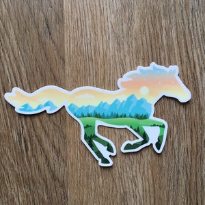May include: A sticker in the shape of a running horse. The horse is filled with a landscape design featuring mountains, trees, and a sunset. The sticker is white with a glossy finish and is placed on a wooden surface.