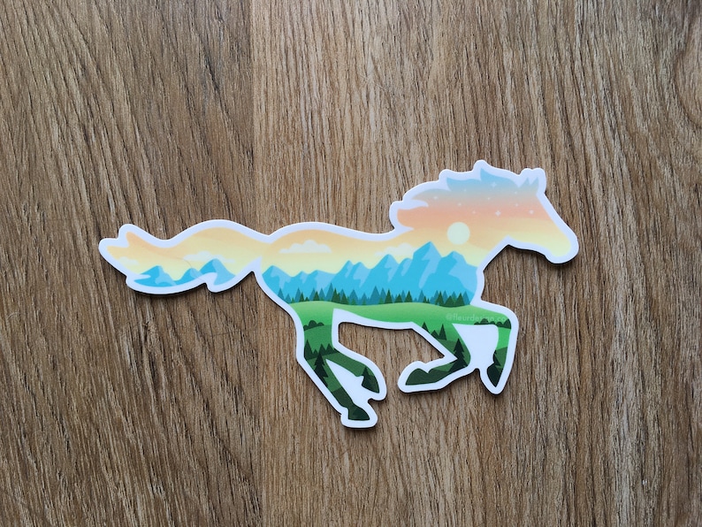 May include: A sticker in the shape of a running horse. The horse is filled with a landscape design featuring mountains, trees, and a sunset. The sticker is white with a glossy finish and is placed on a wooden surface.