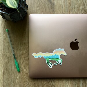 May include: A sticker of a running horse with a landscape design, featuring a green forest, blue mountains, and a yellow and orange sky. The sticker is on a closed laptop. A green pen and a potted plant are also visible.