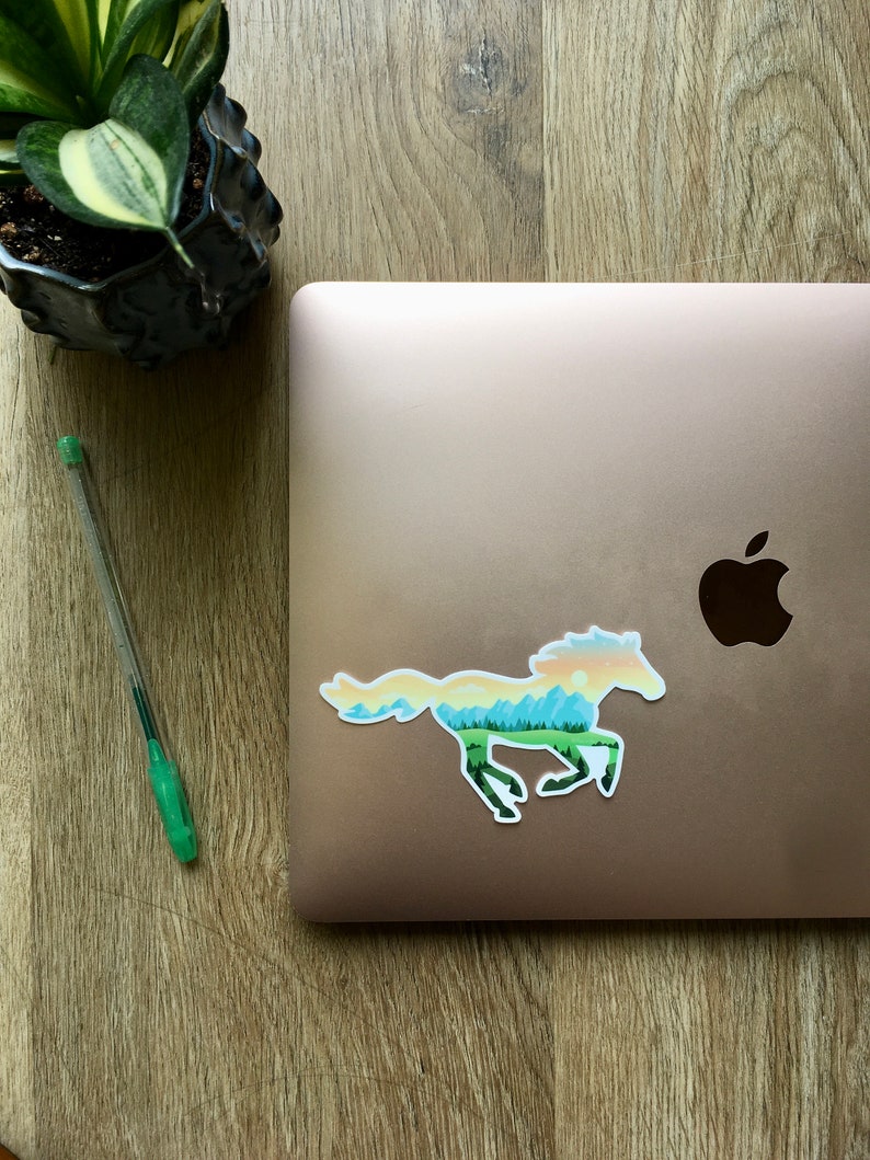 May include: A sticker of a running horse with a landscape design, featuring a green forest, blue mountains, and a yellow and orange sky. The sticker is on a closed laptop. A green pen and a potted plant are also visible.