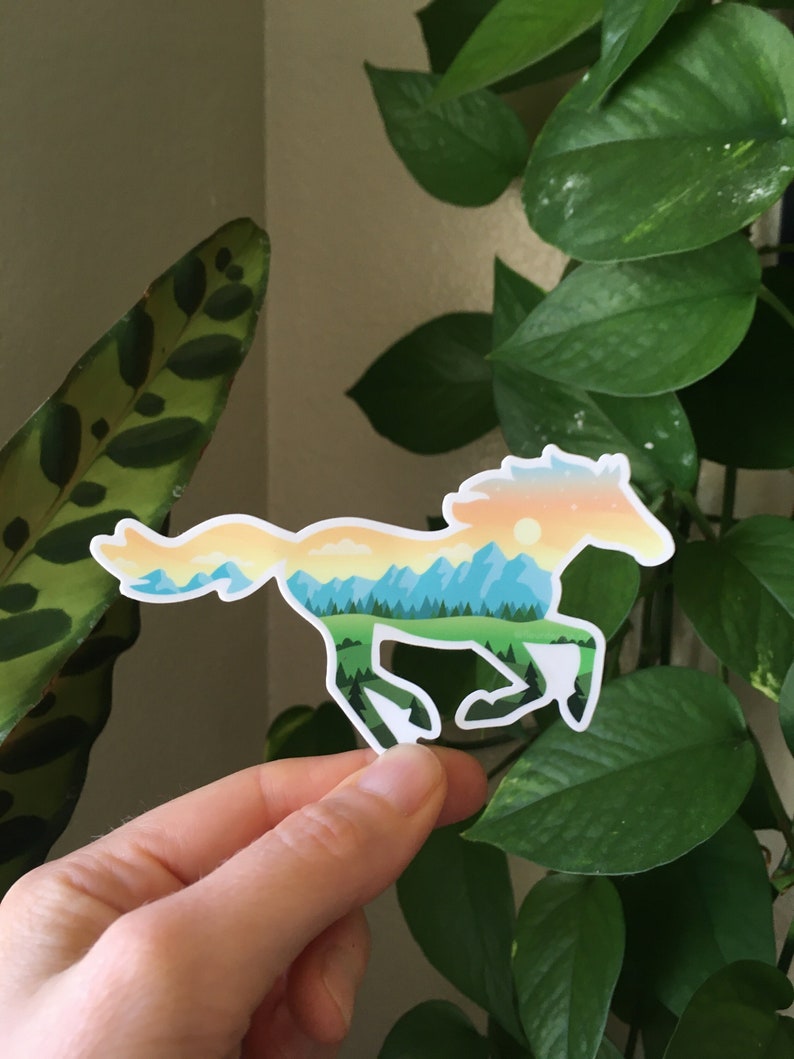 May include: A white sticker depicting a running horse. The horse's outline is filled with a landscape scene of mountains, trees, and a sunset in blue, green, yellow, and orange. The sticker is held in a person's hand.