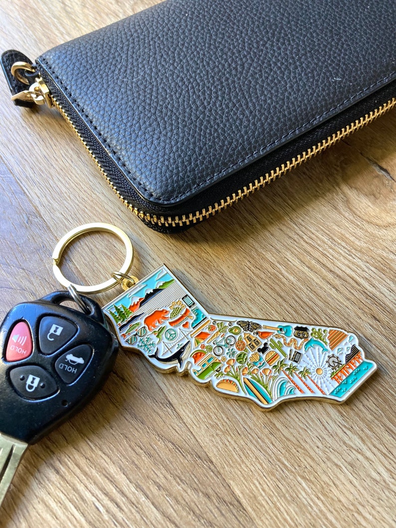 Peut inclure: Un porte-cl&eacute;s en forme de l'&eacute;tat de Californie, de couleur dor&eacute;e, avec un contour d&eacute;taill&eacute; de l'&eacute;tat et diverses ic&ocirc;nes repr&eacute;sentant la culture californienne, notamment des palmiers, des planches de surf et un ours. Le porte-cl&eacute;s est attach&eacute; &agrave; un portefeuille en cuir noir avec une fermeture &eacute;clair dor&eacute;e.