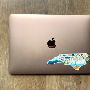 Pu&ograve; includere: Un laptop color oro rosa con un adesivo a forma di stato della Carolina del Nord. L'adesivo &egrave; pieno di icone colorate che rappresentano diversi aspetti dello stato, come montagne, spiagge e citt&agrave;.