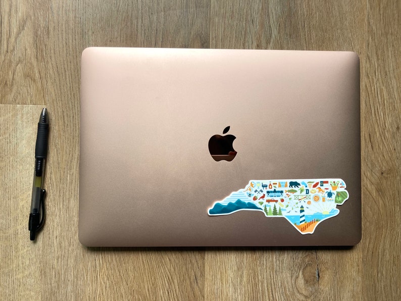 Pu&ograve; includere: Un laptop color oro rosa con un adesivo a forma di stato della Carolina del Nord. L'adesivo &egrave; pieno di icone colorate che rappresentano diversi aspetti dello stato, come montagne, spiagge e citt&agrave;.