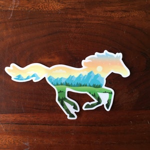May include: A sticker in the shape of a running horse, with a landscape design inside. The horse silhouette is filled with a scene of mountains, trees, and a sunset. The sticker has a white border and is on a wooden background.