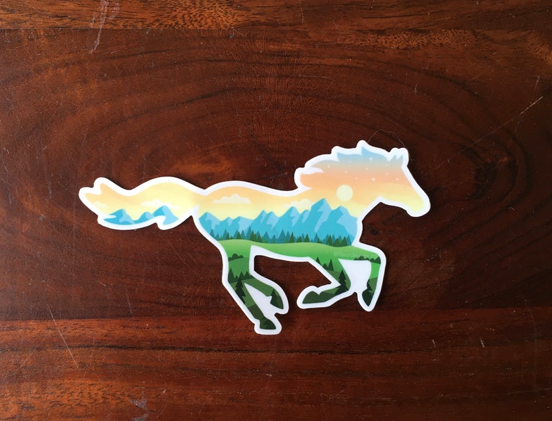 May include: A sticker in the shape of a running horse, with a landscape design inside. The horse silhouette is filled with a scene of mountains, trees, and a sunset. The sticker has a white border and is on a wooden background.