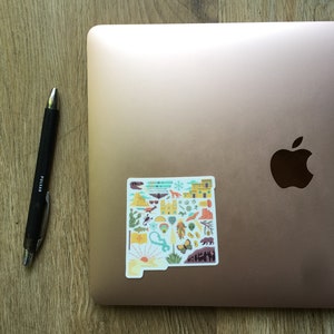 Op de afbeelding: Een sticker van de staat New Mexico met verschillende iconen die de cultuur en het landschap van de staat vertegenwoordigen. De sticker zit op een roze gouden laptop.