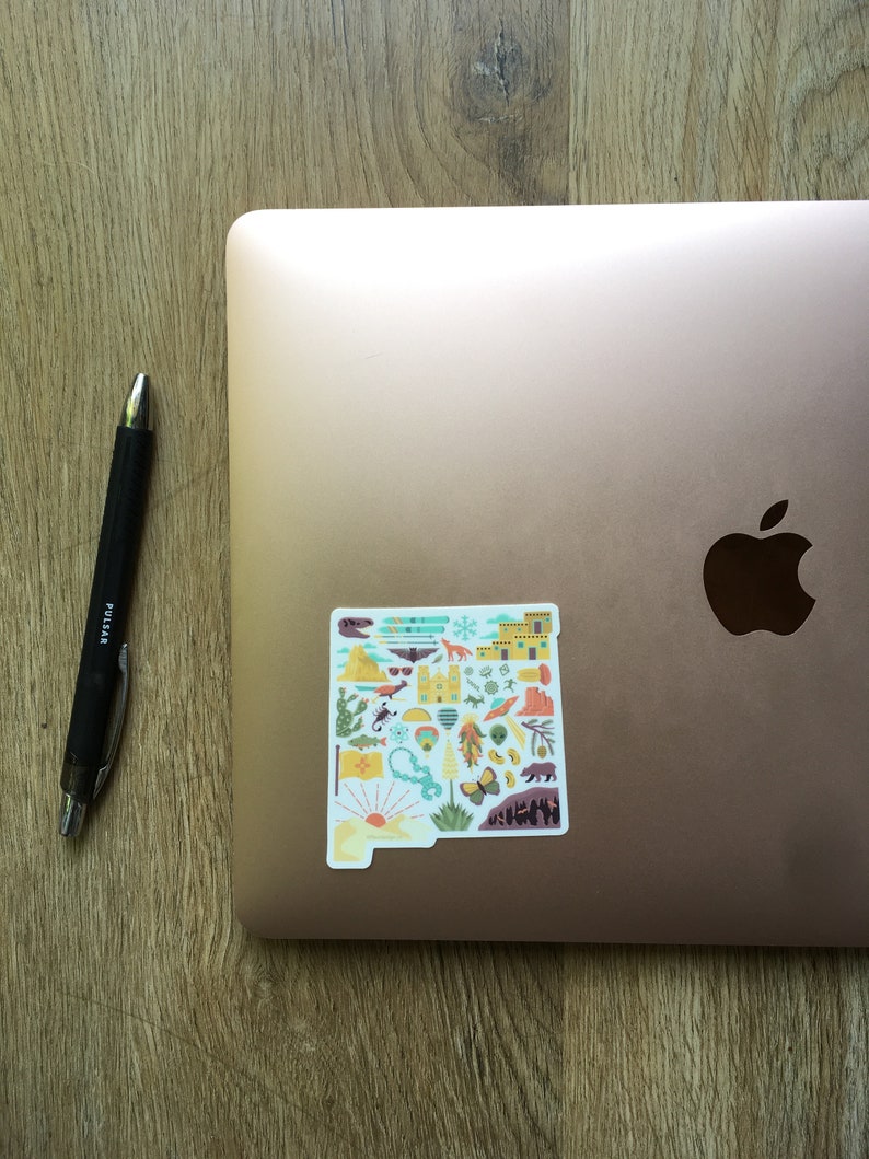 Op de afbeelding: Een sticker van de staat New Mexico met verschillende iconen die de cultuur en het landschap van de staat vertegenwoordigen. De sticker zit op een roze gouden laptop.