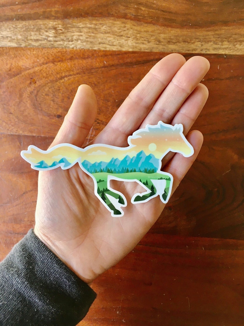 May include: A sticker in the shape of a running horse, with a landscape design. The horse silhouette is filled with a mountain scene in shades of green, blue, yellow, and orange. The sticker is held in a hand.