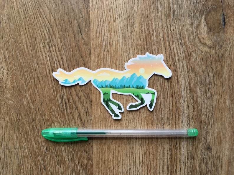 May include: A sticker in the shape of a running horse, with a landscape design inside. The landscape features mountains, trees, and a sunset. A green pen is below the sticker on a wooden surface.