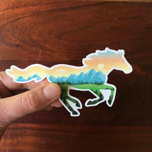 May include: A sticker in the shape of a running horse, with a landscape design. The horse silhouette is filled with a scene of mountains, trees, and a sunset in shades of blue, green, yellow, and orange. The sticker is held by a hand.