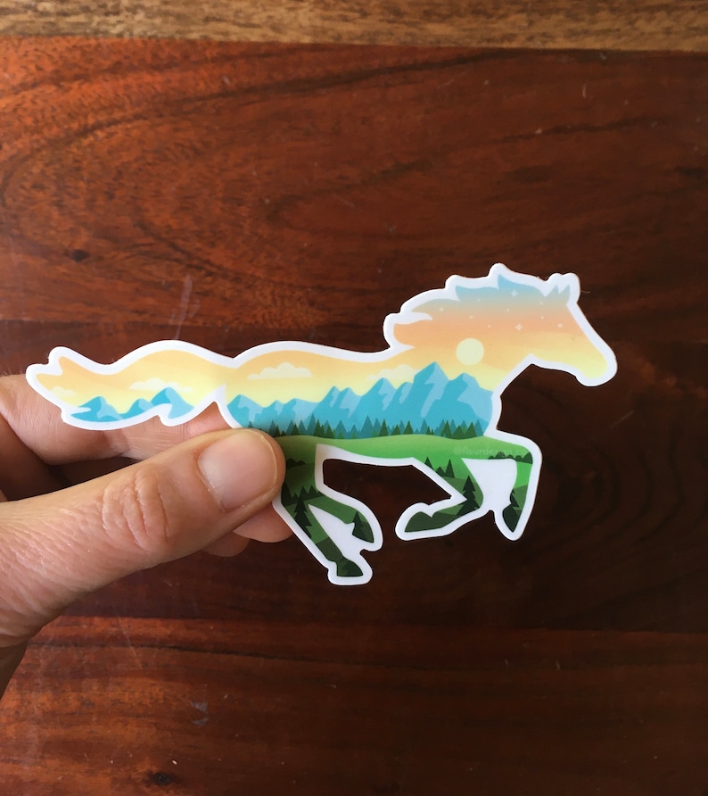May include: A sticker in the shape of a running horse, with a landscape design. The horse silhouette is filled with a scene of mountains, trees, and a sunset in shades of blue, green, yellow, and orange. The sticker is held by a hand.