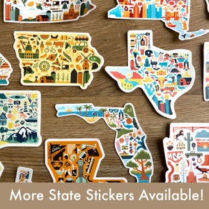 Pu&ograve; includere: Una collezione di adesivi colorati a forma di stato con illustrazioni di monumenti e simboli. Gli adesivi sono su una superficie di legno. Il testo "More State Stickers Available!" &egrave; nella parte inferiore dell'immagine.