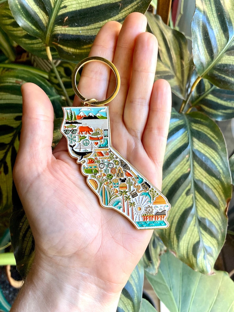 Peut inclure: Porte-cl&eacute;s en m&eacute;tal dor&eacute; en forme de l'&eacute;tat de Californie, avec un fond en &eacute;mail blanc et des illustrations color&eacute;es de monuments californiens, notamment un ours, des palmiers, une plage et une planche de surf.
