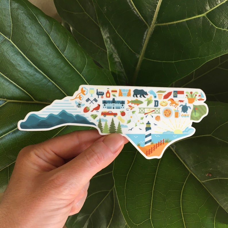Pu&ograve; includere: Un adesivo bianco a forma dello stato della Carolina del Nord con illustrazioni colorate di monumenti, animali e attivit&agrave;. L'adesivo include il testo "OBX".