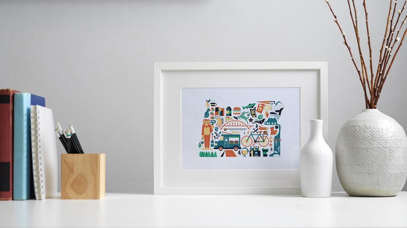 A framed Oregon map art print is leaning against a wall. The art consists of a collection illustrated icons that form the shape of the state of Oregon. Each illustration represents that culture, natural beauty and history of the state of Oregon.