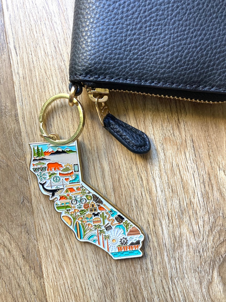 Peut inclure: Un porte-cl&eacute;s en forme de l'&eacute;tat de Californie, de couleur dor&eacute;e, avec une illustration d&eacute;taill&eacute;e de diverses ic&ocirc;nes repr&eacute;sentant la culture californienne, notamment des palmiers, des planches de surf et une for&ecirc;t de s&eacute;quoias. Le porte-cl&eacute;s est attach&eacute; &agrave; un portefeuille en cuir noir avec une fermeture &eacute;clair dor&eacute;e.