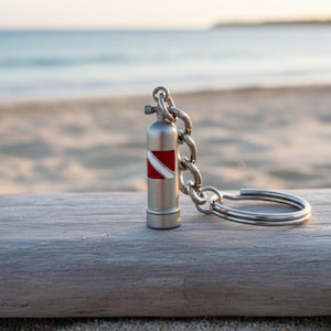 May include: A silver-colored scuba tank keychain with a red and white dive flag design. The keychain is attached to a silver chain and a split ring. The background shows a beach and ocean.