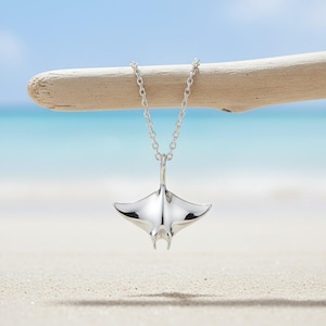 May include: A silver-colored pendant necklace featuring a manta ray design. The pendant is suspended from a delicate chain and is displayed against a beach backdrop with a blue sky and driftwood.