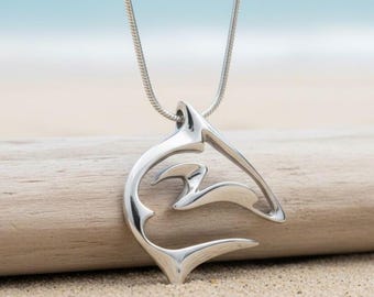 May include: A silver-colored pendant necklace featuring a stylized wave design. The pendant is suspended from a thin, silver chain. The necklace is displayed on a piece of driftwood on a sandy beach, with the ocean in the background.