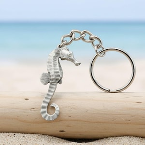 May include: A silver-toned metal keychain featuring a detailed seahorse charm attached to a circular split ring by a short chain. The seahorse has a textured, segmented body with a curved tail and a small fin, finished in a matte metallic sheen.