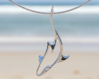 May include: A silver pendant necklace featuring a stylized shark design. The pendant is suspended from a thin, silver chain. The shark pendant has a smooth, polished finish and is set against a blurred beach background.