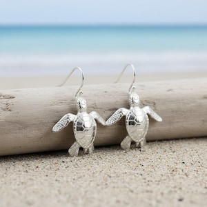 May include: A pair of silver-colored turtle earrings. Each earring features a detailed turtle design with outstretched flippers and a textured shell. The earrings hang from simple, curved ear wires. The background shows a sandy beach and ocean.