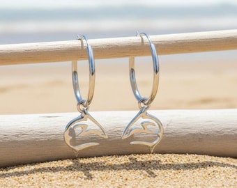 May include: A pair of silver hoop earrings with a dolphin tail design. The earrings are suspended from a wooden dowel against a backdrop of a sandy beach and ocean. The earrings are a bright, shiny silver color.