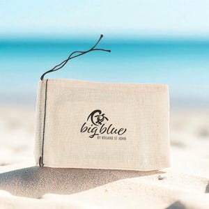May include: A rectangular, off-white canvas bag with the words "big blue" and a turtle logo printed in black. The bag is tied with a black string and sits on a sandy beach with a turquoise ocean in the background.