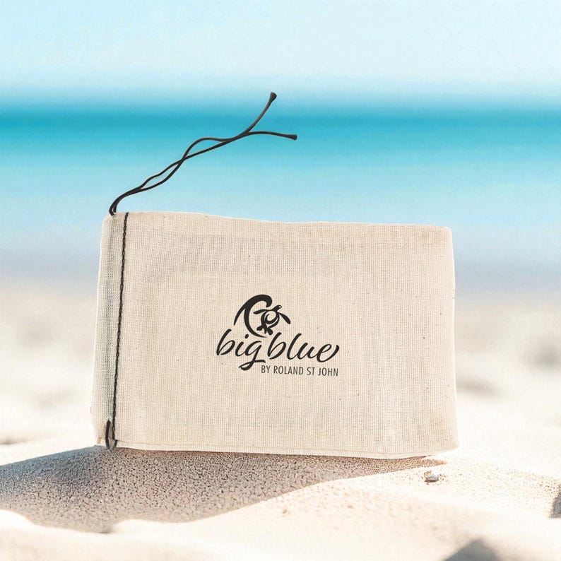 May include: A rectangular, off-white canvas bag with the words "big blue" and a turtle logo printed in black. The bag is tied with a black string and sits on a sandy beach with a turquoise ocean in the background.