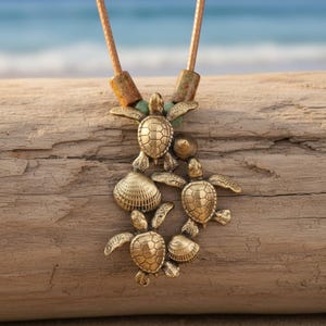 May include: A necklace featuring a brass-toned pendant with three sea turtles and two seashells. The pendant is suspended from a tan cord with two textured, cylindrical beads at the top, showcasing intricate shell and turtle scale details.