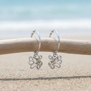 May include: A pair of silver hoop earrings with octopus charms. The earrings are displayed on a piece of driftwood against a backdrop of sand and ocean. The octopus charms have detailed tentacles.