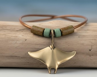 Könnte beinhalten: Eine Halskette mit einem metallisch goldfarbenen Mantarochen-Anhänger an einer dünnen braunen Kordel. Die Kordel ist mit zwei zylindrischen braunen Perlen und zwei kleineren hellgrünen Perlen auf beiden Seiten der Anhängeröse verziert.