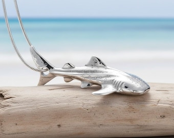 May include: A silver shark pendant on a silver chain. The shark pendant is detailed with fins, gills, and a textured surface. The necklace is displayed on a piece of driftwood with a blurred ocean background.