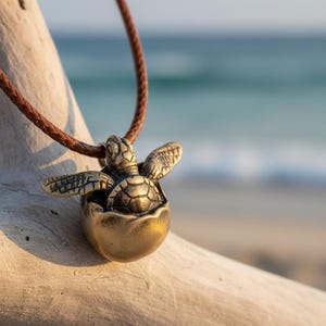 May include: A bronze-colored turtle pendant necklace. The turtle is emerging from an egg-shaped shell, suspended from a brown braided leather cord. The background shows a blurred beach and ocean scene.