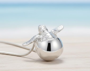 May include: A silver turtle pendant necklace. The turtle is perched atop a spherical base, with its flippers extended. The necklace chain is a thin, silver snake chain. The background is a blurred beach scene with a light blue sky and sandy shore.