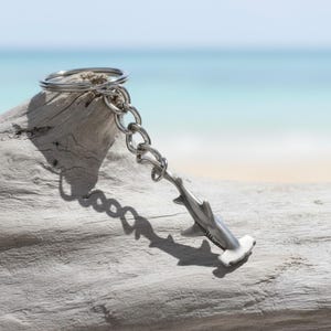 May include: A silver-toned metal keychain featuring a hammerhead shark charm attached to a circular split ring by a short chain. The shark charm has a smooth, matte finish and detailed fins, suspended from a standard metal keyring component.