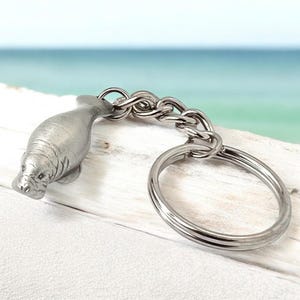 May include: A silver-toned metal keychain featuring a three-dimensional manatee charm attached to a sturdy link chain and a circular split ring. The manatee figure displays detailed textures on its body, including visible facial features and fins.
