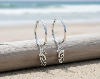 May include: A pair of silver hoop earrings with seahorse charms. The earrings are displayed on a piece of driftwood on a sandy beach, with the ocean in the background. The seahorse charms are detailed and dangle from the hoops.