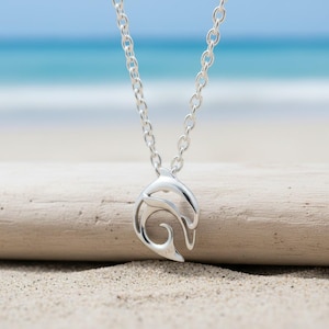 May include: A silver-colored pendant necklace featuring a stylized dolphin design. The pendant is suspended from a delicate chain. The background shows a sandy beach and the ocean.